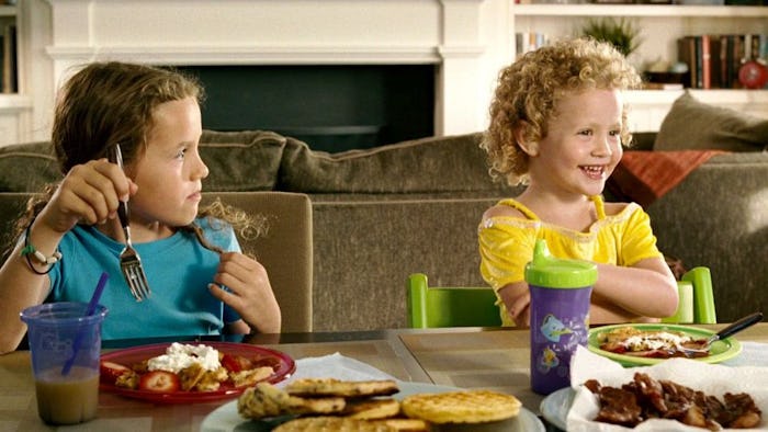 Two preschoolers sitting at a dining table; one of them is eating and the other one is smiling with ...
