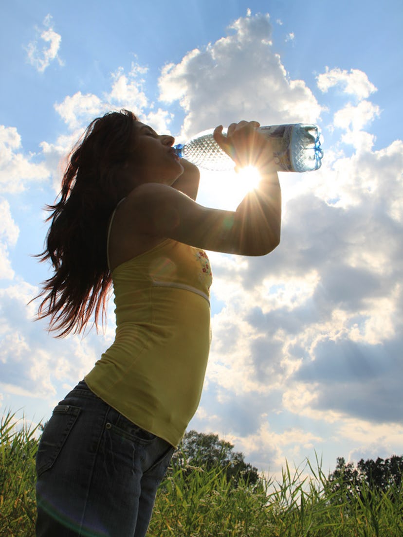 A woman drinking water from a bottle with the sun shining behind the clouds in her background.