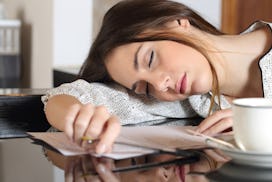 An exhausted woman sleeping on a pile of papers while holding a pen with a cup of coffee on a table ...
