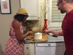 A woman and a man standing in their kitchen both holding knives and slicing a cake
