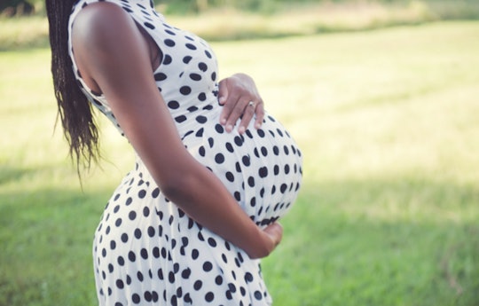 A pregnant woman standing in a white dress with black dots and holding her stomach with her hands