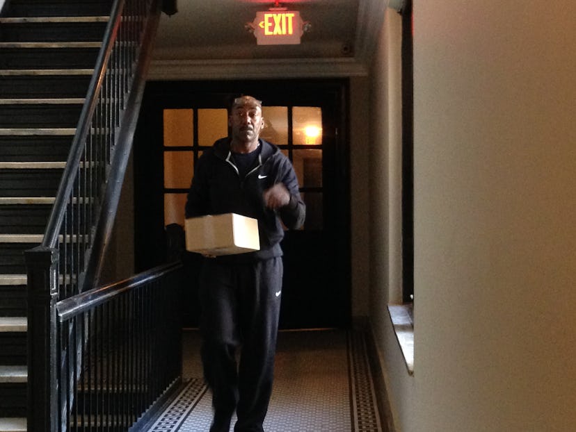 A man dressed in a black tracksuit walking in a building hallway carrying a delivery box.