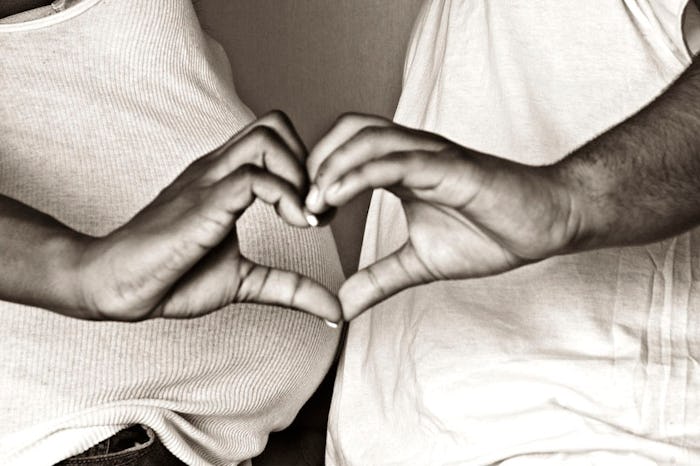 black and white photo of a pregnant woman and partner standing belly to belly, making a heart with t...
