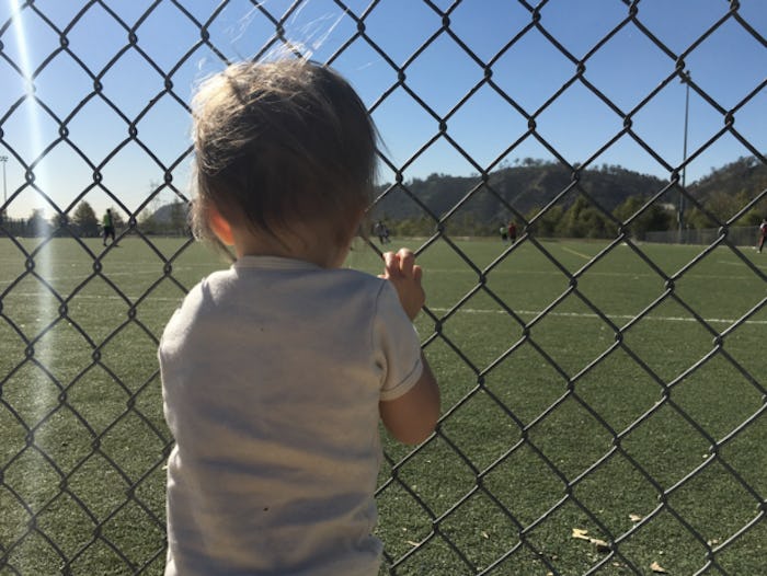 A little kid holding on to the fence of a field