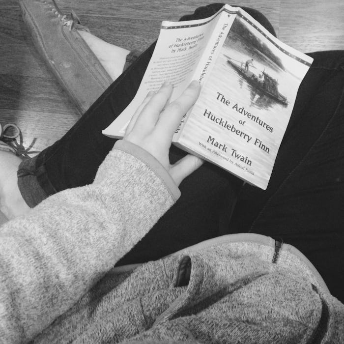 A black and white photo of a person holding a copy of The Adventures of Huckleberry Finn on their la...