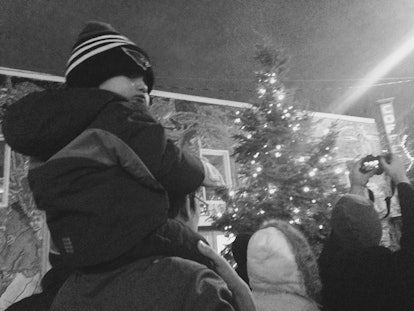 A man is carrying a little boy on his back outside in front of a big Christmas tree.