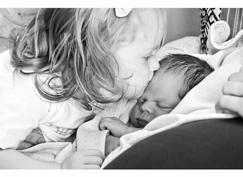 A little girl with short and curly hair is kissing the newborn's forehead.