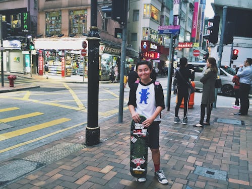 A kid growing up in Hong Kong holding his skateboard while standing on the street