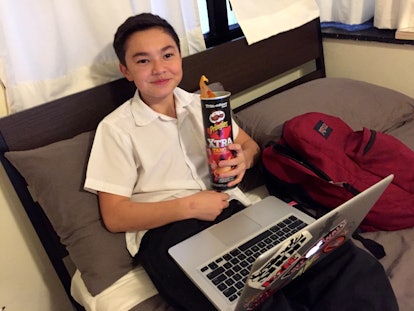 A tween boy sitting on a couch with a laptop in his lap, holding a package of chips