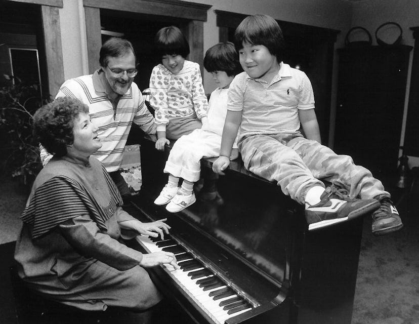 Lacey sitting on the piano with her siblings. The mother is playing the piano, and the dad is standi...