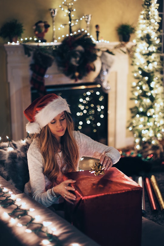 A woman sitting on a couch in a santa hat with christmas decorations behind her opening a large red ...