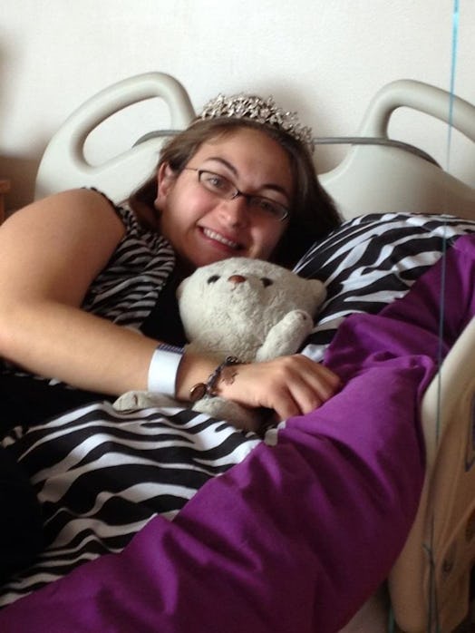 Photo of Megan Zander lying in a hospital bed, wearing a crown on her head, hugging her teddy, and s...