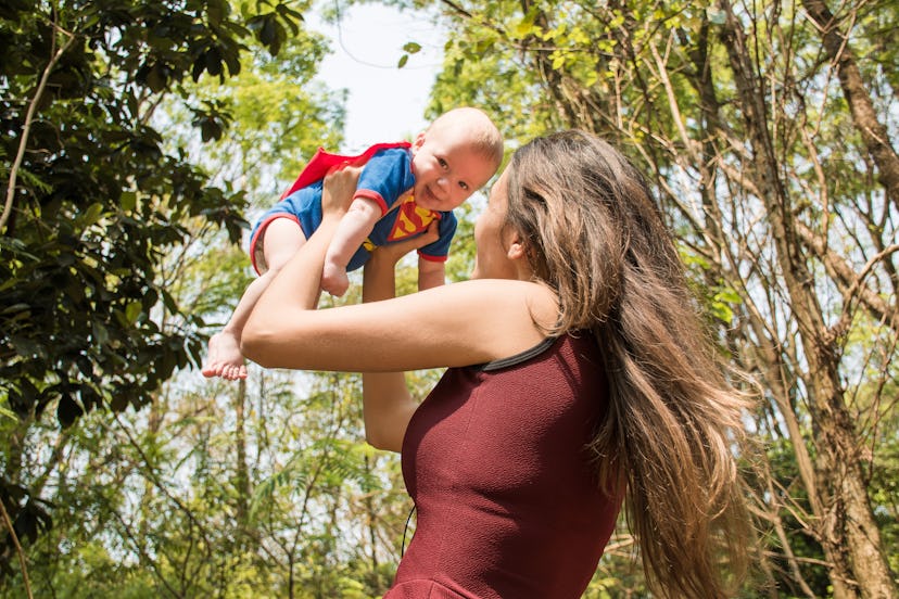 A mother holding her happy baby dressed in a Superman costume in the air in a park.
