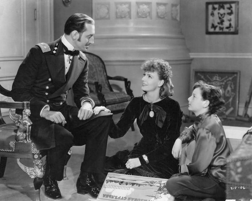 Black and white image of a man wearing a suite and sitting on a chair, talking to two women