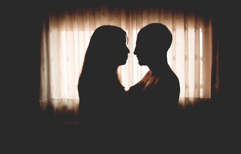 A couple standing facing each other in a dark room with a window in front of them