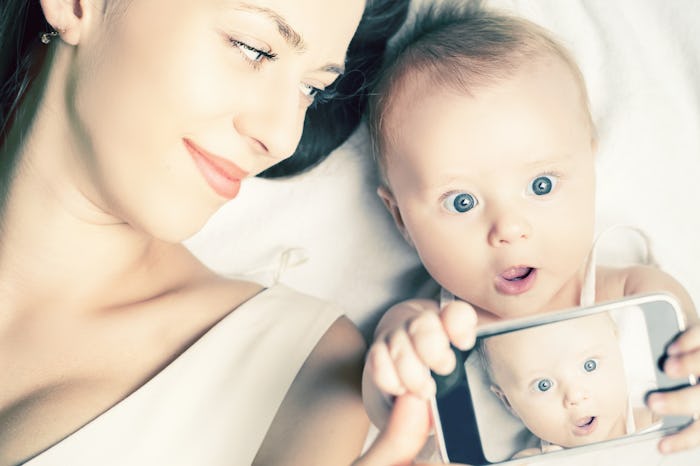 Mom lying with her baby in bed, while baby is holding a phone and taking a selfie