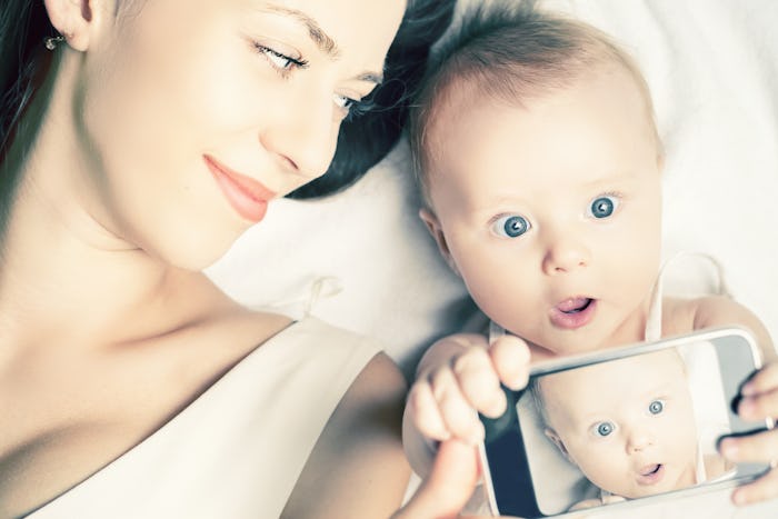 Mom lying with her baby in bed, while baby is holding a phone and taking a selfie