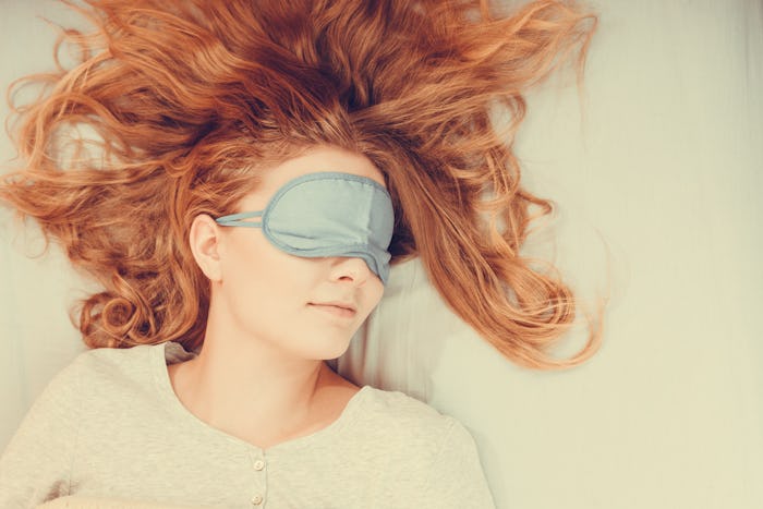 A red-haired woman in a white top and a grey sleep mask, sleeping on her back