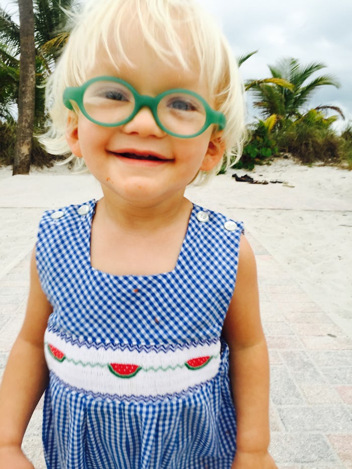 A blonde kid in green glasses and a blue romper with watermelons on it 