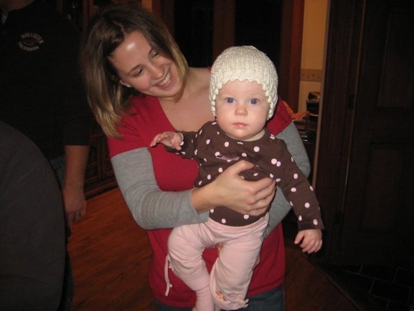 A woman wearing a red shirt smiles while holding a baby wearing a white knitted hat and brow shirt w...