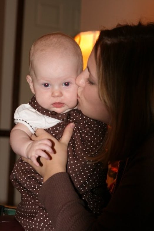 A woman holding a baby wearing a polka dot dress in her arms and kissing the baby's cheek.