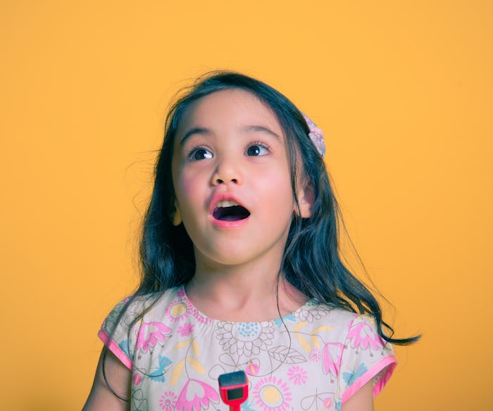 A little girl in a pink dress standing in front of a yellow background with her mouth open