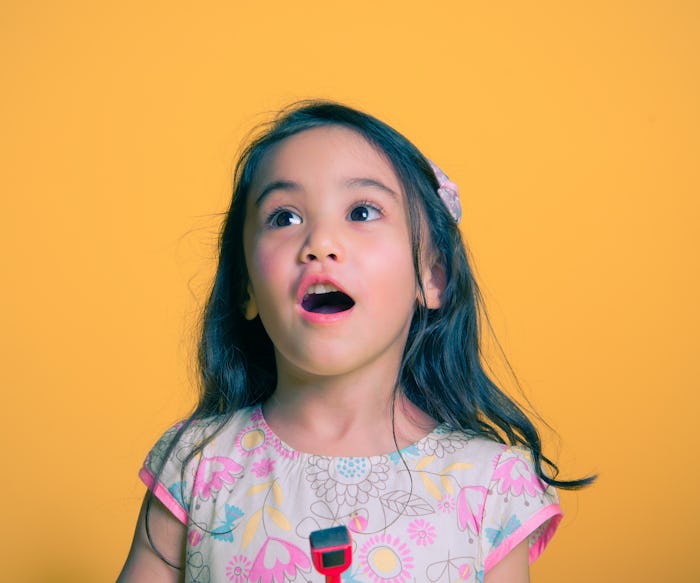 A little girl in a pink dress standing in front of a yellow background with her mouth open