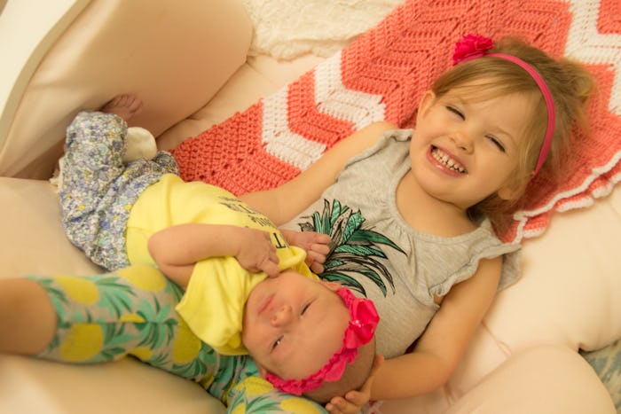 A toddler holding her newborn baby sister while sitting together on the couch