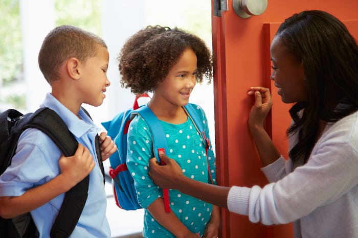 Two children wearing backpacks in the hall at school talk to a teacher who is crouching next to them