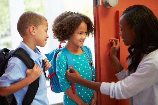 Two children wearing backpacks in the hall at school talk to a teacher who is crouching next to them