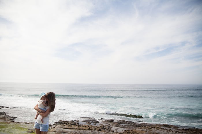 A new mom holding her baby on a beach next to the sea.