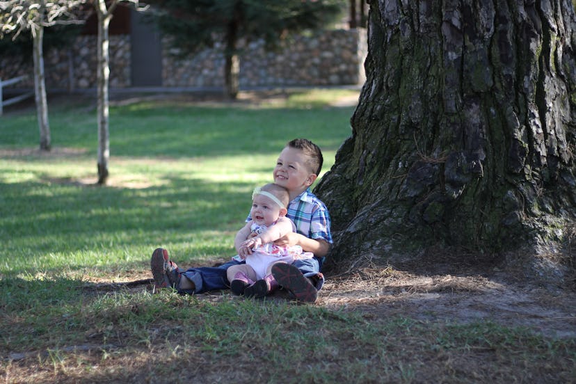 An older brother is holding his baby sister while they are sitting on the grass