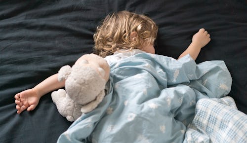 A blonde child lying stomach-down in its bed with a plush toy during bedtime