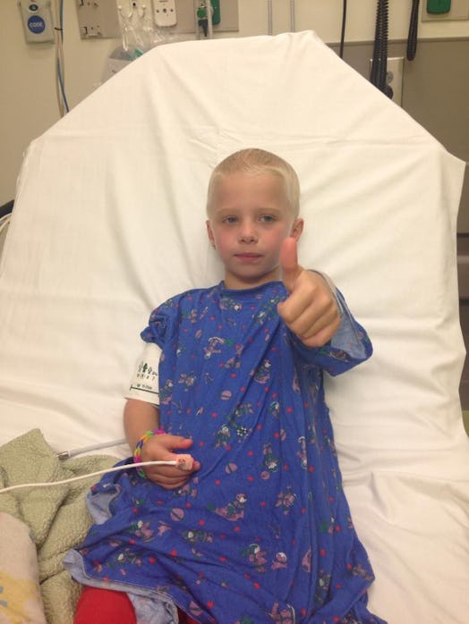 A girl with Leukemia with a shaved head sitting in a hospital bed and showing a thumb up