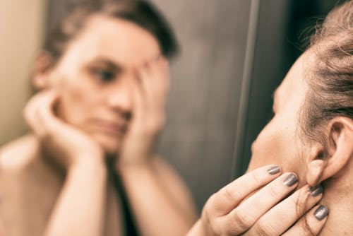 A woman with Postpartum Depression looks at herself in the mirror touching her face and neck.