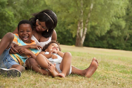 A mother and her two children sitting on the ground in the park, she's hugging them while they laugh...