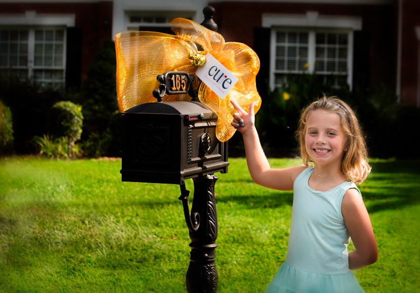 The girl who has Leukemia smiling and pointing at a mail box with a large orange ribbon and a card w...