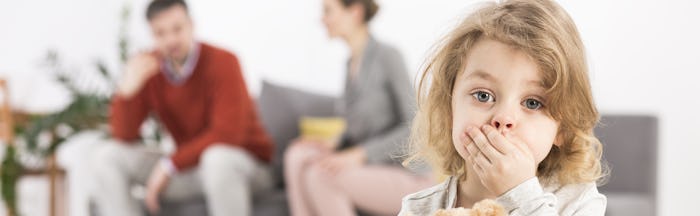 A toddler covering her mouth while her parents are blurred in the background