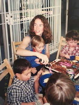 A mother sitting with four kids at the table and cutting the cake while one of them is sitting in he...
