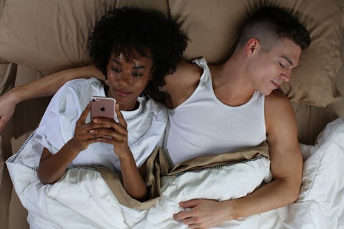 A man sleeping in a bed and a woman lying next to him typing on her phone