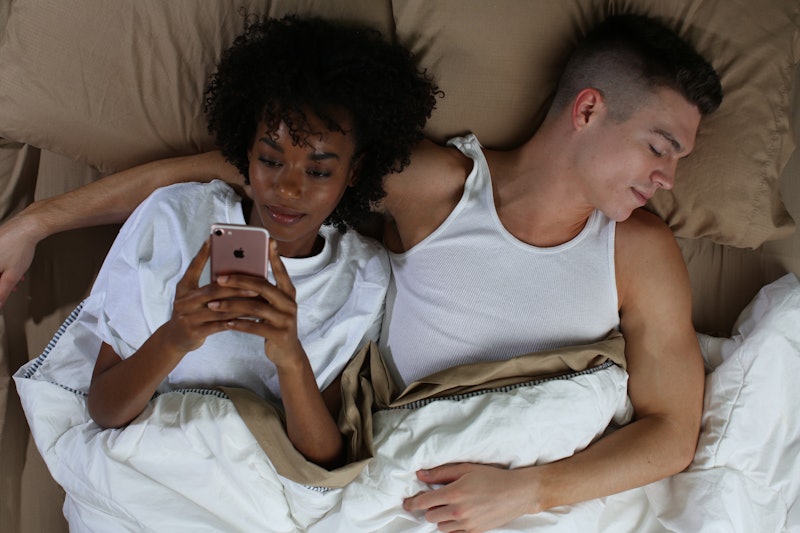 A man sleeping in a bed and a woman lying next to him typing on her phone