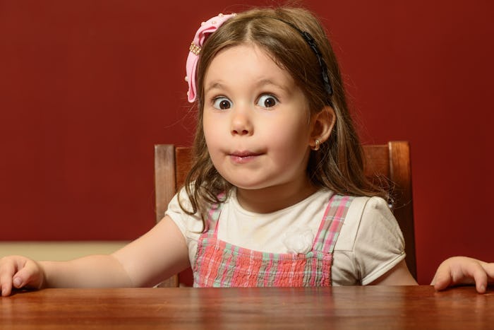 A little girl with a pink bow in her hair sitting at the desk with a surprised look 