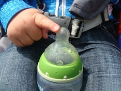 A toddler sitting in a car seat with a baby bottle full of soy milk in his lap