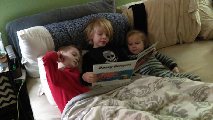 Three kids lying in a bed and the one in the middle is reading a book