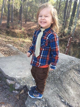 A little boy wearing brown pants and a checked shirt with a tie, smiling