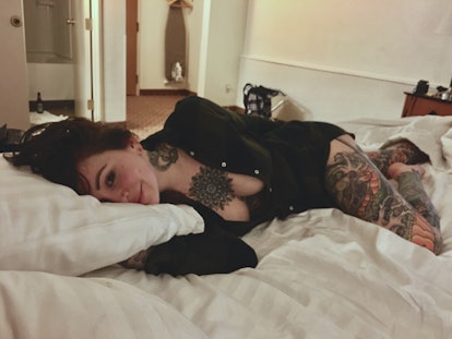 A tattooed woman wearing a black unbuttoned shirt is lying provocatively on a bed and looking at the...