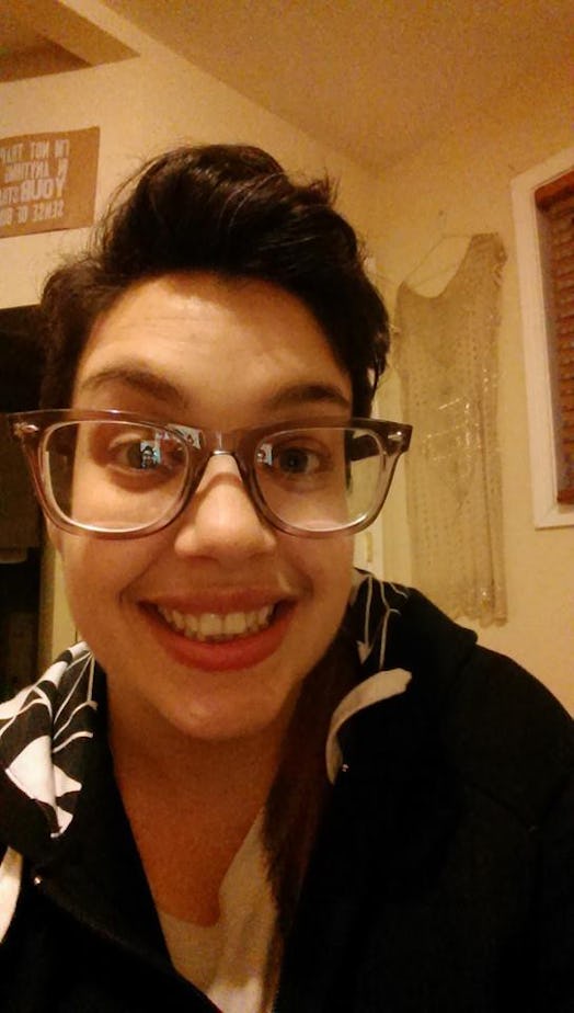A non-binary transgender person wearing glasses is taking a selfie while smiling.