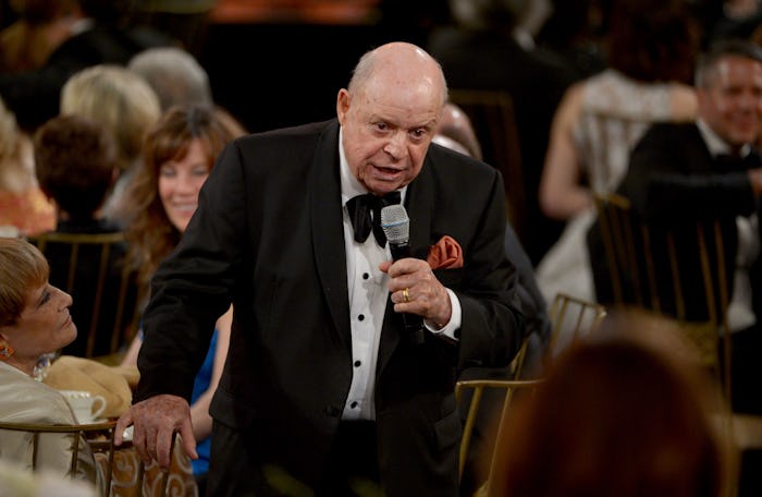 Don Rickles in a black suit, holding a microphone and speaking