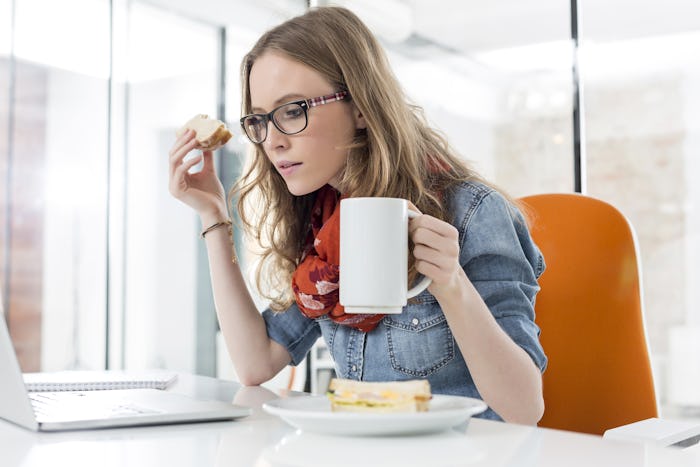 A girl holding a cup of coffee and piece of sandwich while looking at her laptop