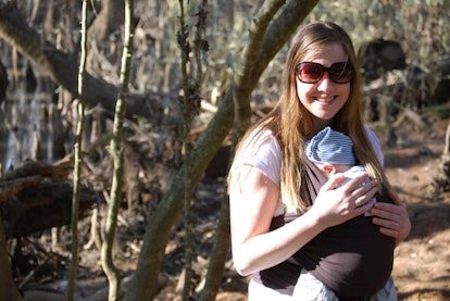 Woman carrying her child in a sling standing in the woods on a sunny day.